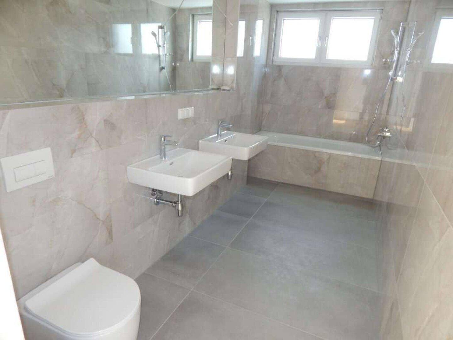 Modernes Badezimmer mit doppelten Waschbecken, Badewanne und großen grauen Fliesen.