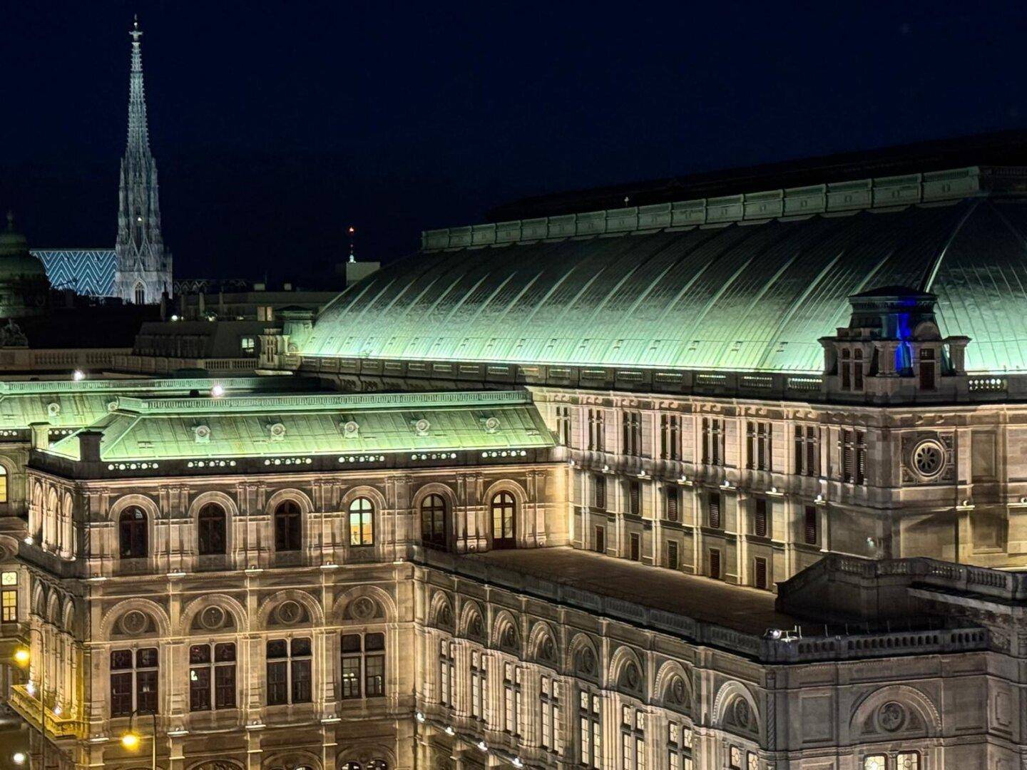 Nächtliche Ansicht eines historischen Gebäudes mit beleuchteter Fassade und einem Kirchturm im Hintergrund.