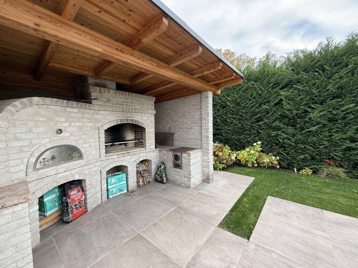 Überdachte Außenküche mit Grill, Pizzaofen und Steinmauern im Gartenbereich.