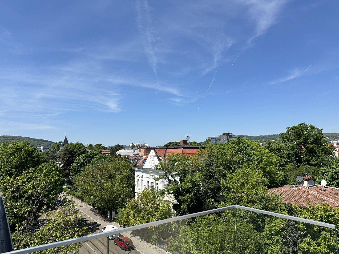 Panoramablick über die Stadtlandschaft mit viel Grün und fernen Hügeln unter blauem Himmel.