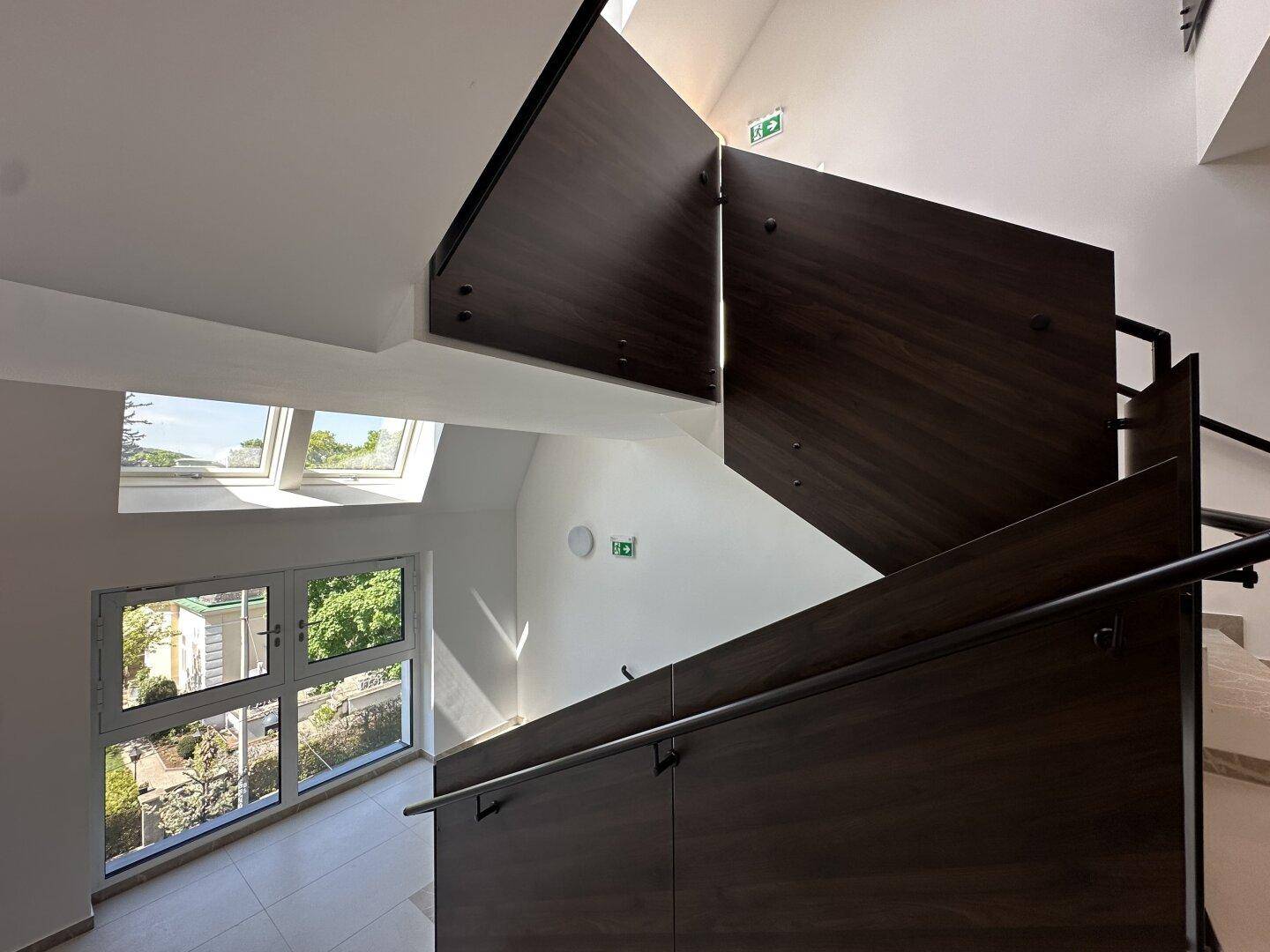 Elegantes Treppenhaus mit dunklen Holzpaneelen, Metallgeländer und viel Tageslicht durch Fenster.