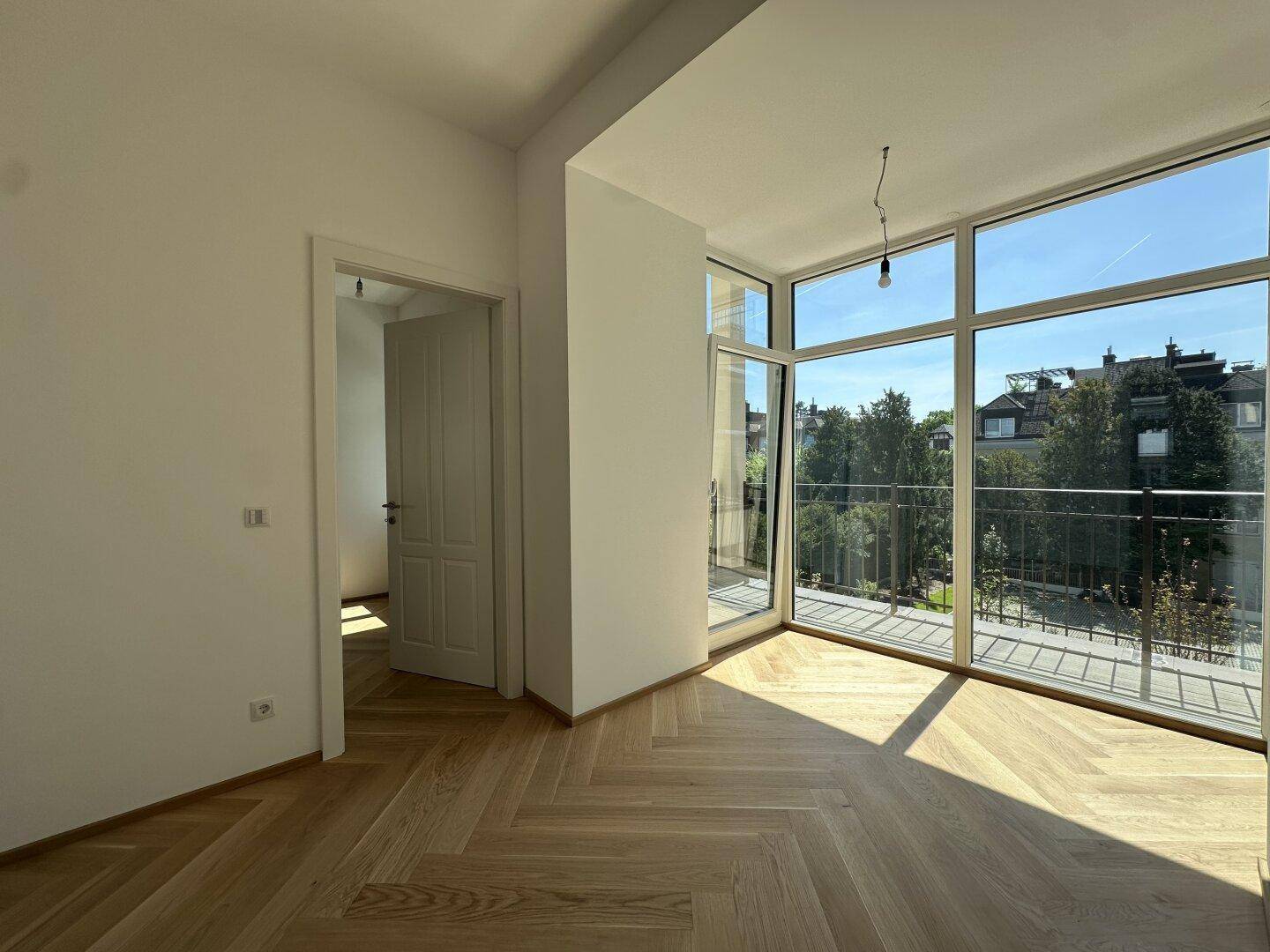 Helles Zimmer mit Fischgrätparkettboden, großen Fenstern und Zugang zu einem französischen Balkon.