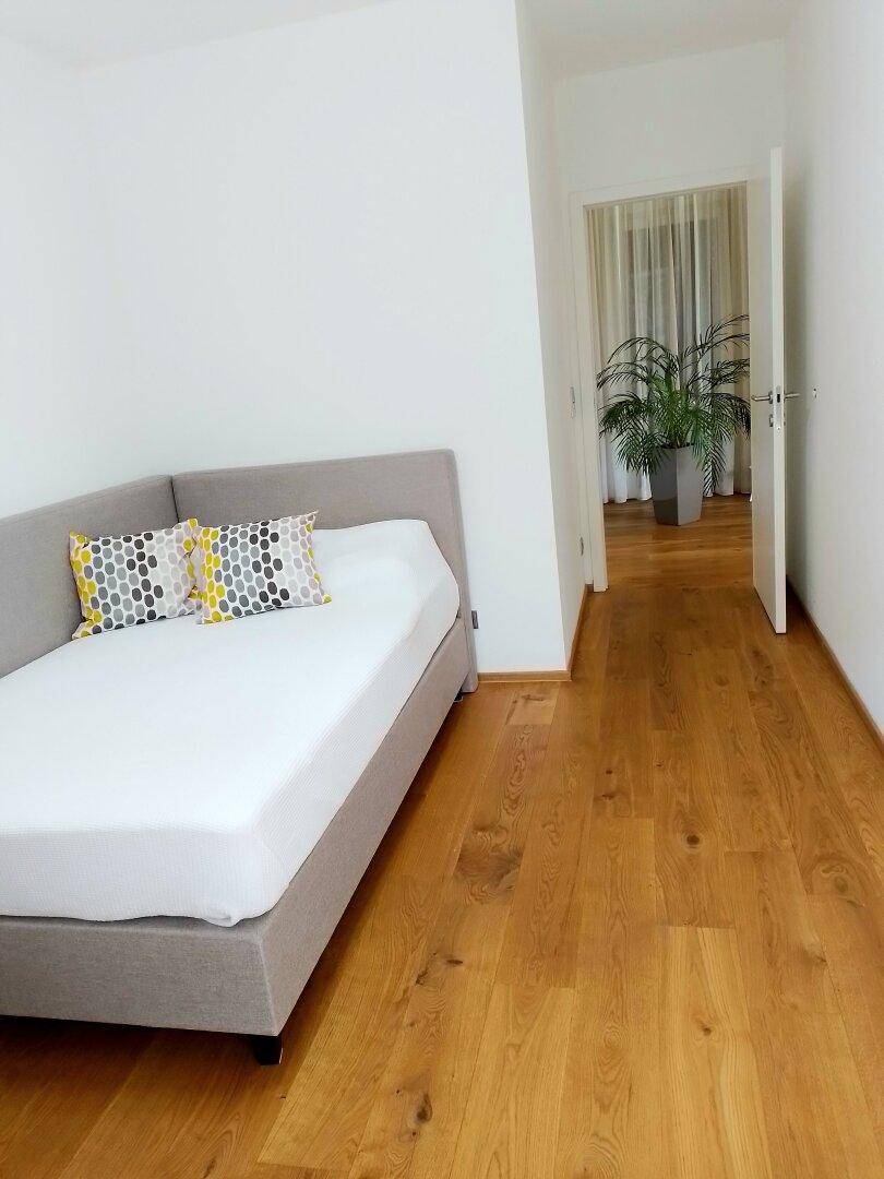 Helles Schlafzimmer mit Einzelbett, Holzboden und Blick in den Flur.