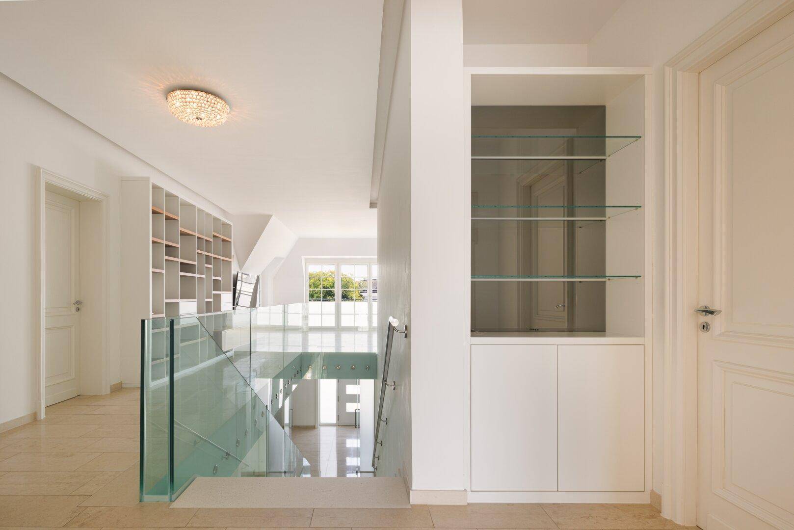Heller Flur im Obergeschoss mit Glasgeländer und Einbauregalen, bietet eine offene und moderne Atmosphäre.