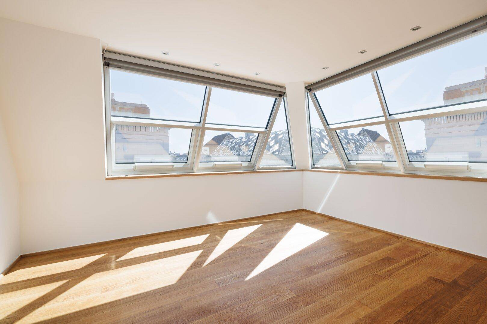 Heller Raum mit großen, schrägen Fenstern und Holzboden, durchflutet von Sonnenlicht.