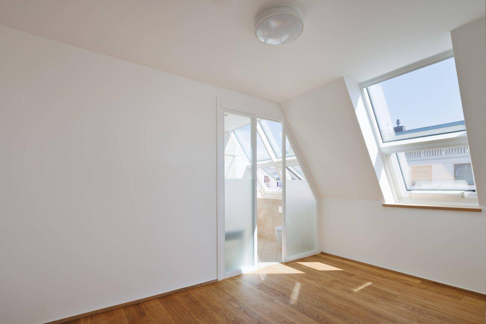 Helles Zimmer mit Holzboden und Dachfenster, mit Blick durch eine Glastür in ein Badezimmer.
