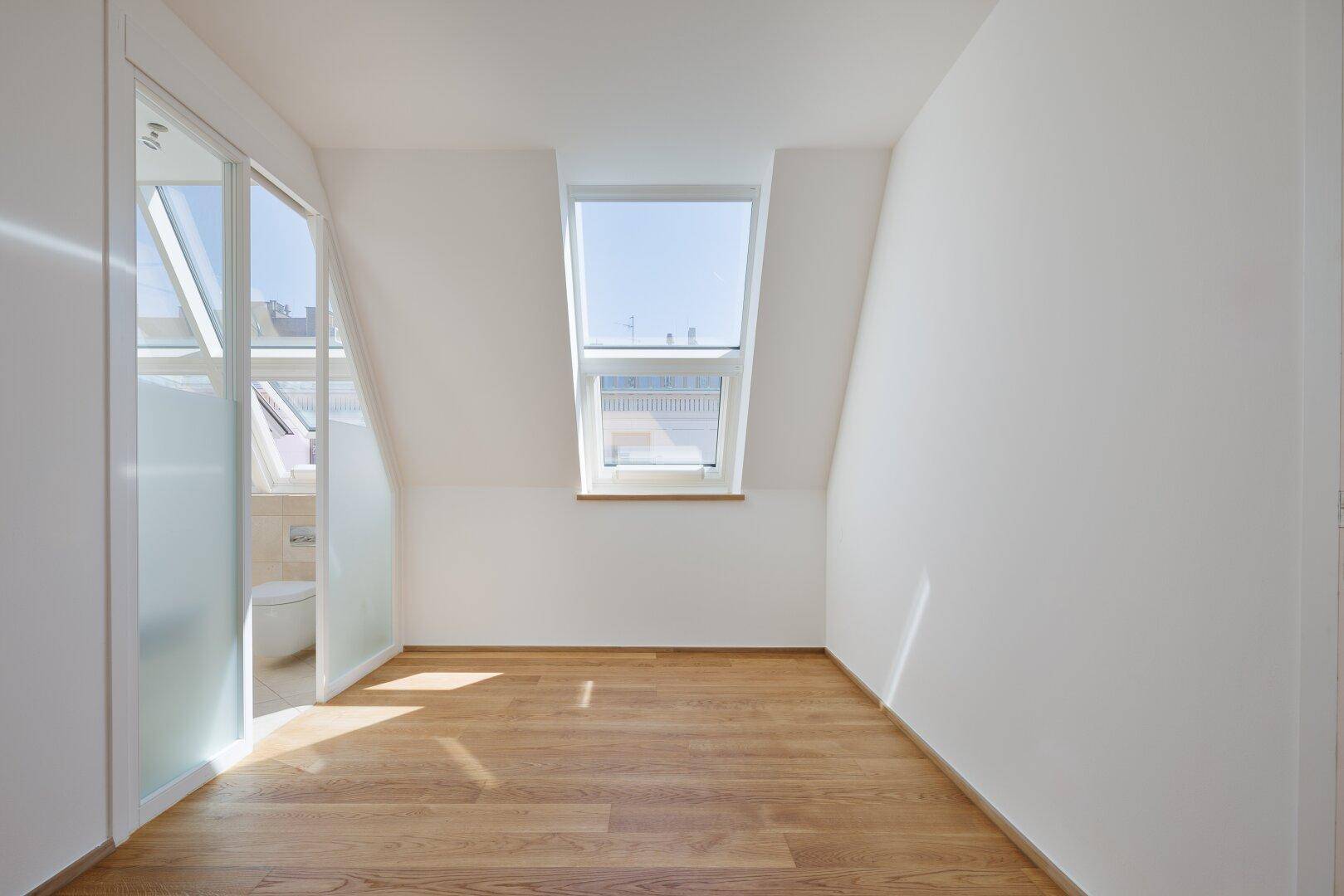 Lichtdurchflutetes Zimmer mit Holzboden und Dachfenster, angrenzend an ein modernes Badezimmer.