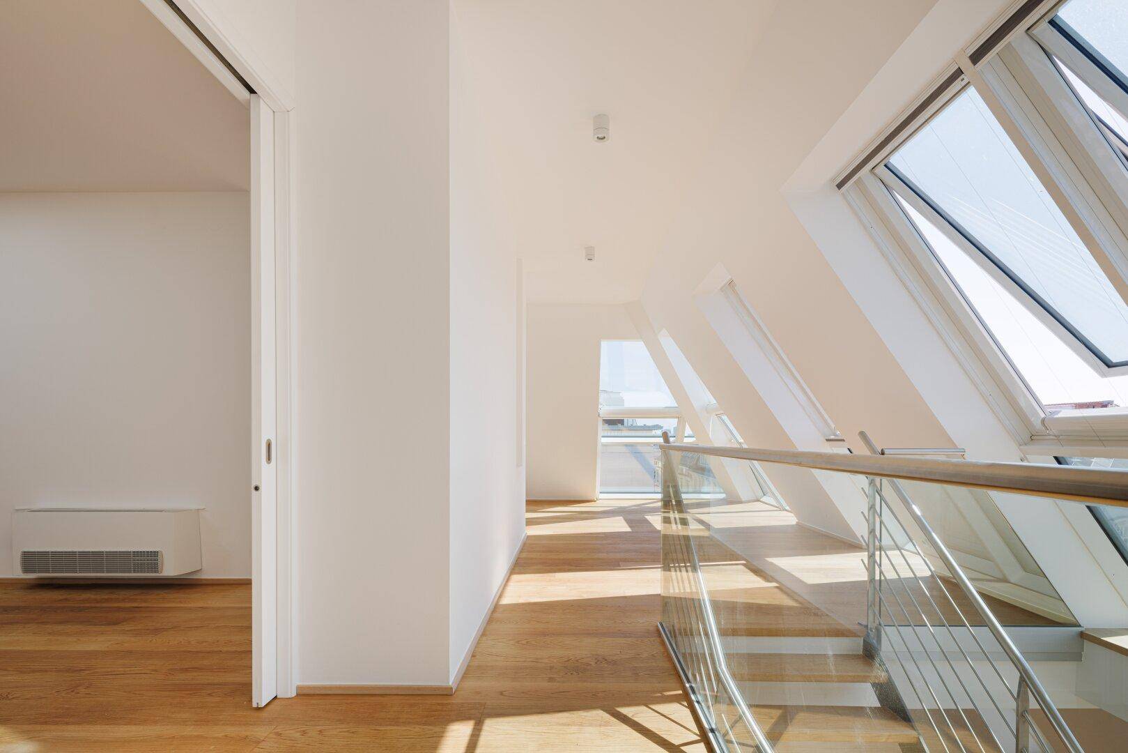 Heller Flur mit Holztreppe, Glasgeländer und schrägen Dachfenstern, die den Raum erhellen.
