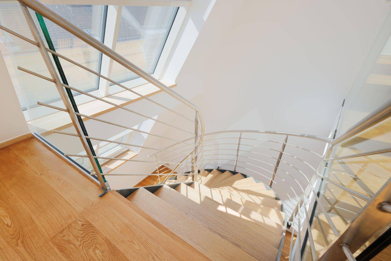 Elegante Holztreppe mit modernem Glas- und Metallgeländer, beleuchtet durch schräge Dachfenster.