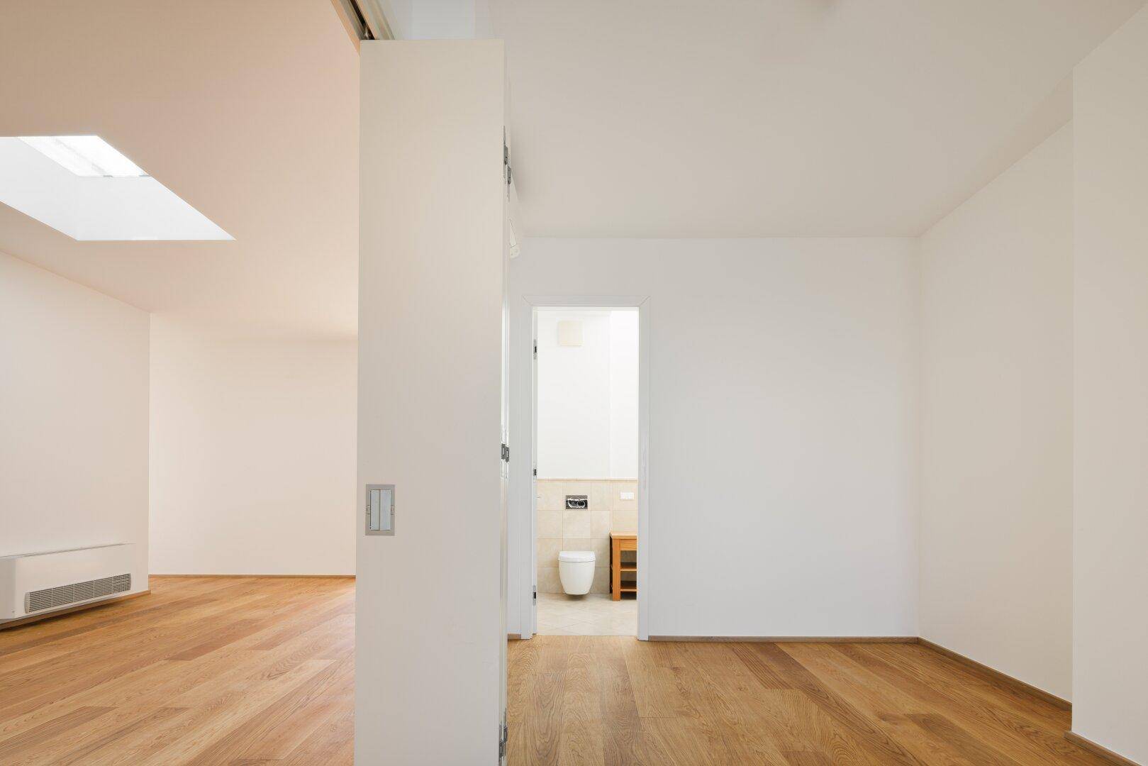 Heller Raum mit Holzboden und Blick in ein Badezimmer mit WC und Waschbecken.