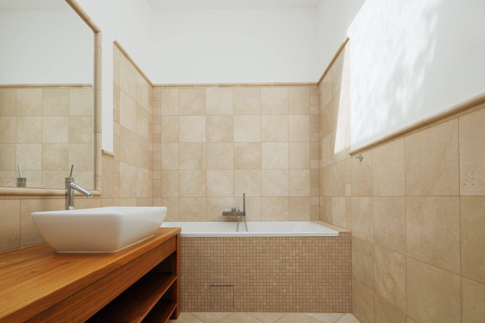 Modernes Badezimmer mit Badewanne, beigen Fliesen und elegantem Waschbecken auf Holzunterschrank.