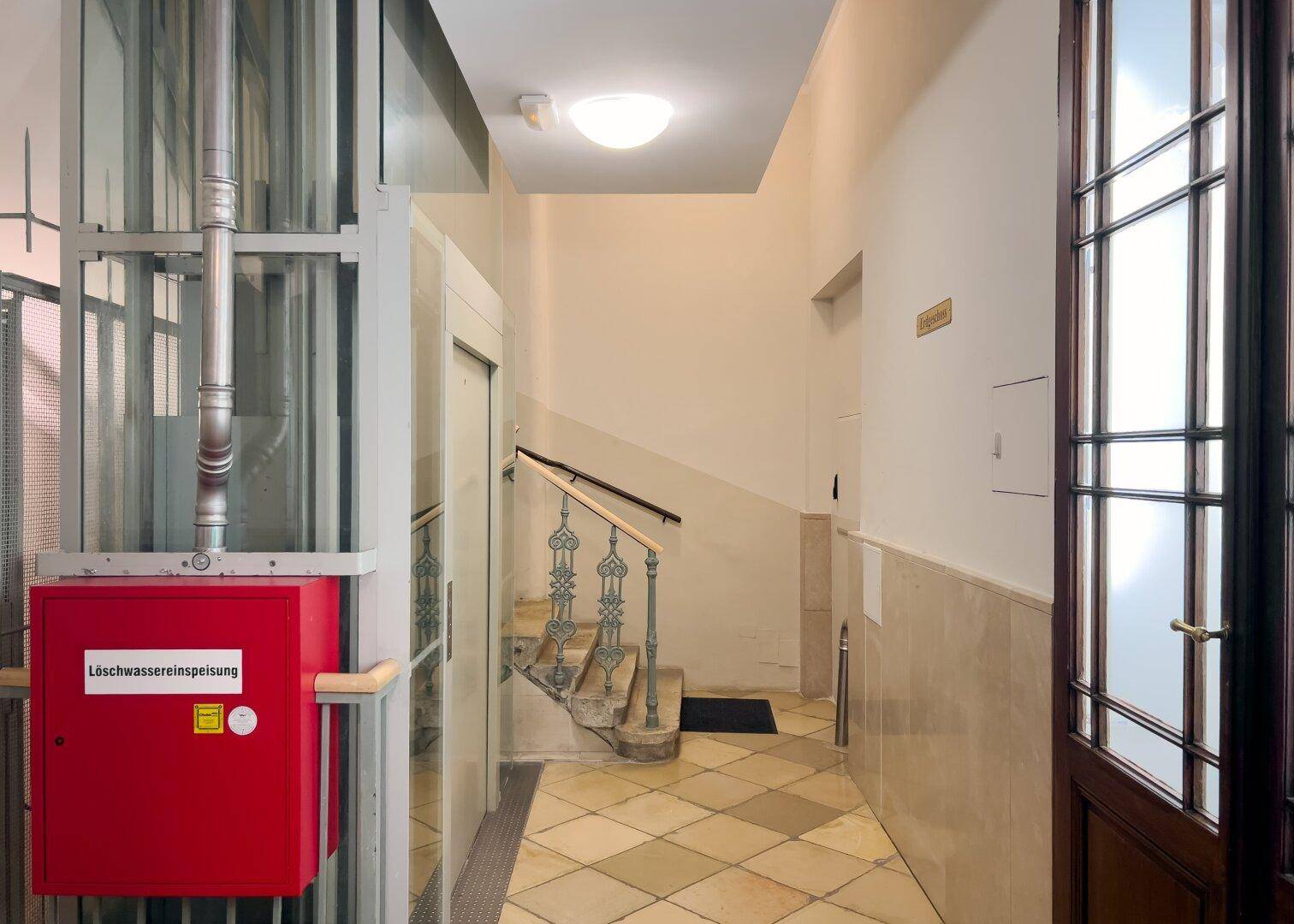 Heller Flur mit Fliesenboden, Treppe und einem modernen Aufzug im Eingangsbereich.