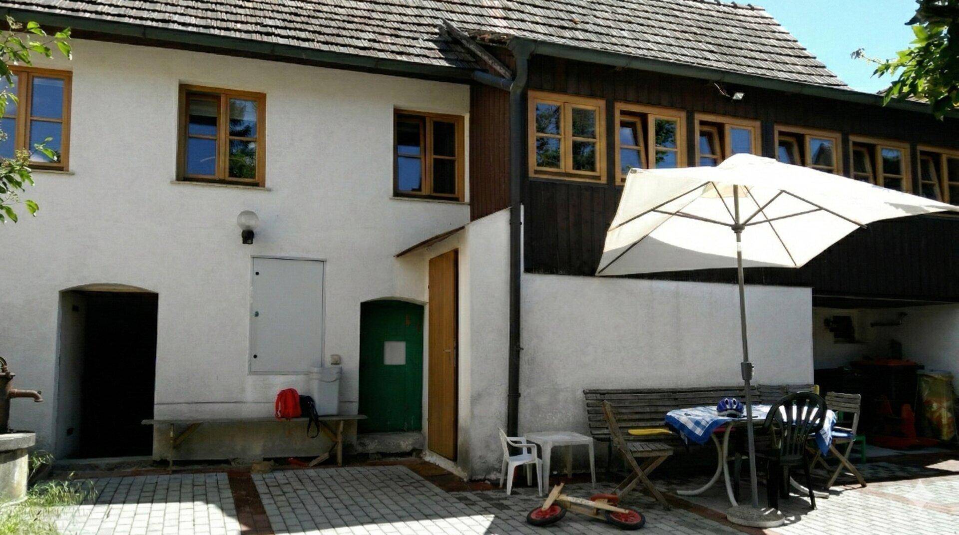 Gemütlicher Außenbereich mit Sitzgelegenheiten und Sonnenschirm vor einem traditionellen Gebäude.