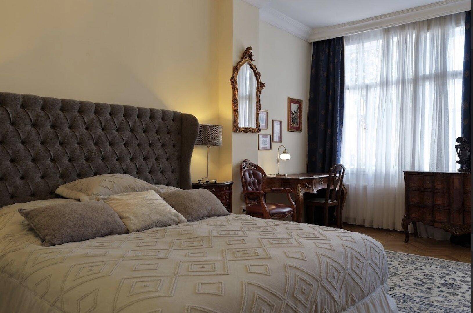 Elegantes Schlafzimmer mit großem gepolstertem Bett, klassischem Schreibtisch und dunklen Vorhängen.