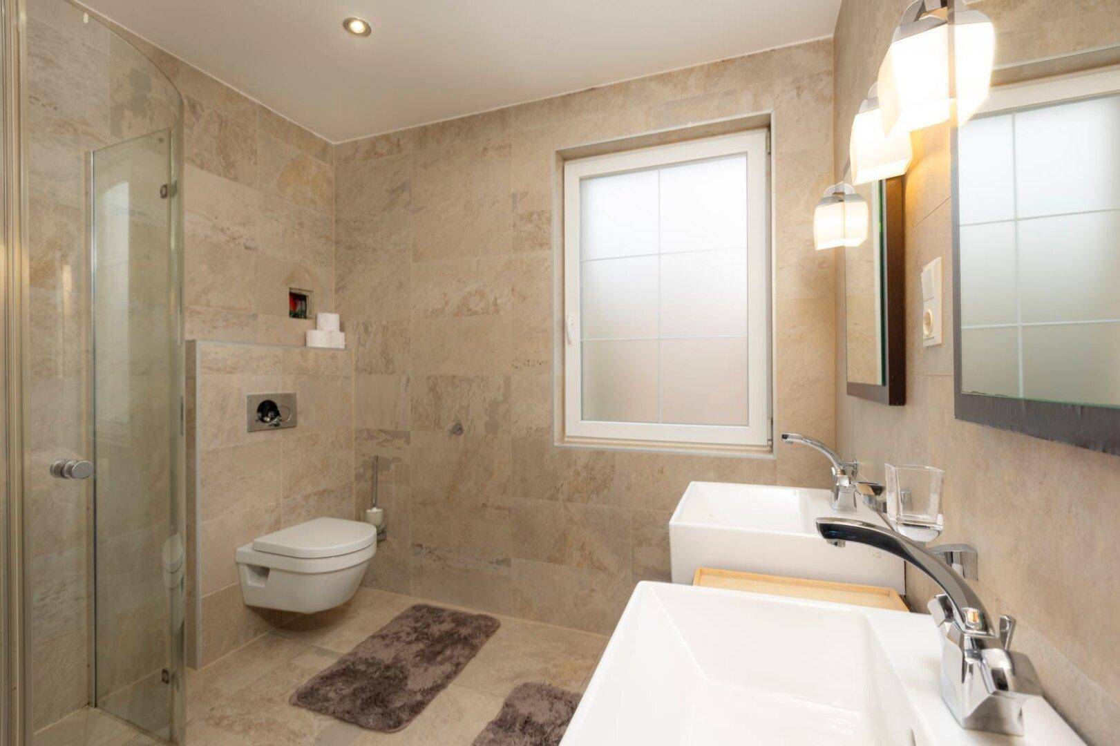 Modernes Badezimmer mit Doppelwaschbecken, Dusche und Fenster, in hellen Fliesen gehalten.