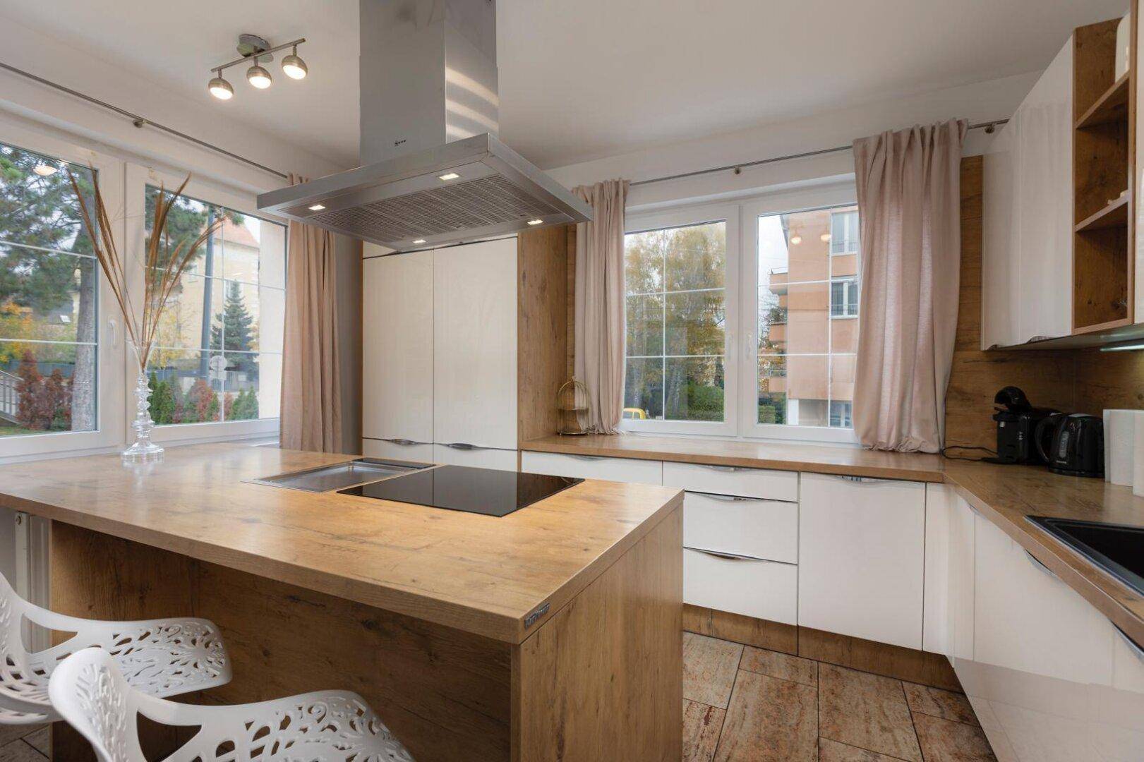 Moderne Küche mit Holz- und weißen Fronten, Kochinsel und großen Fenstern mit Blick ins Grüne.