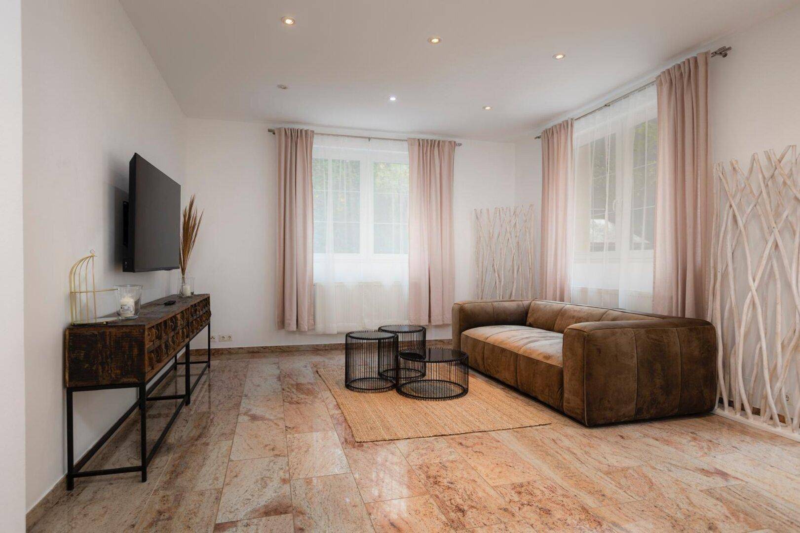 Geräumiges Wohnzimmer mit braunem Sofa, Couchtischen und Fernseher, auf glänzenden Fliesen.