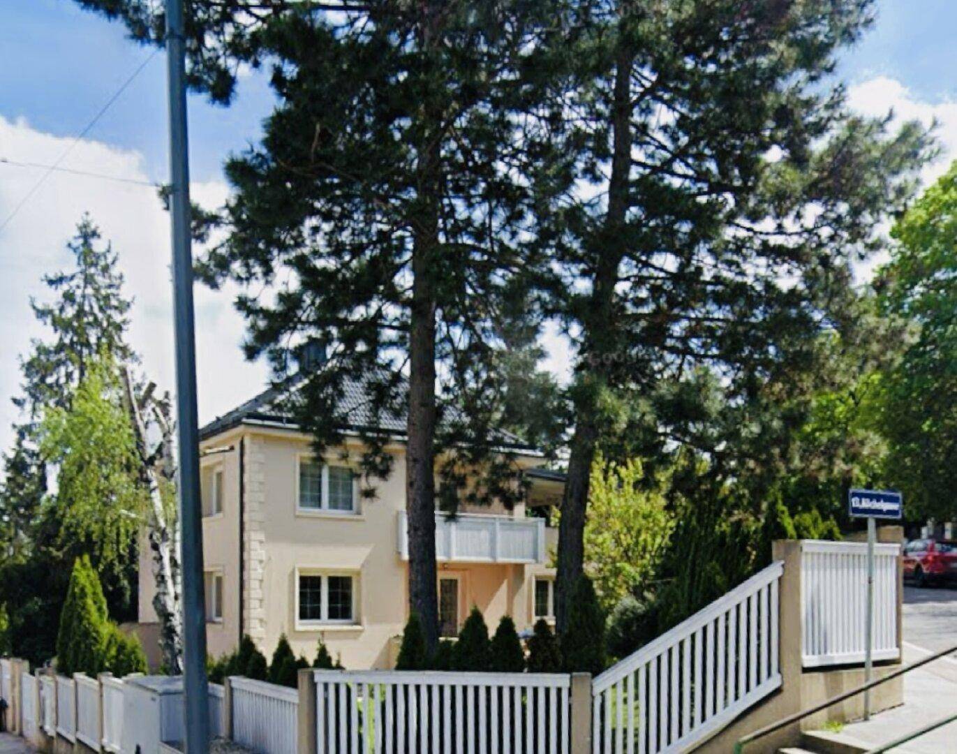 Einfamilienhaus von der Straße aus gesehen, umgeben von Bäumen und einem weißen Zaun.
