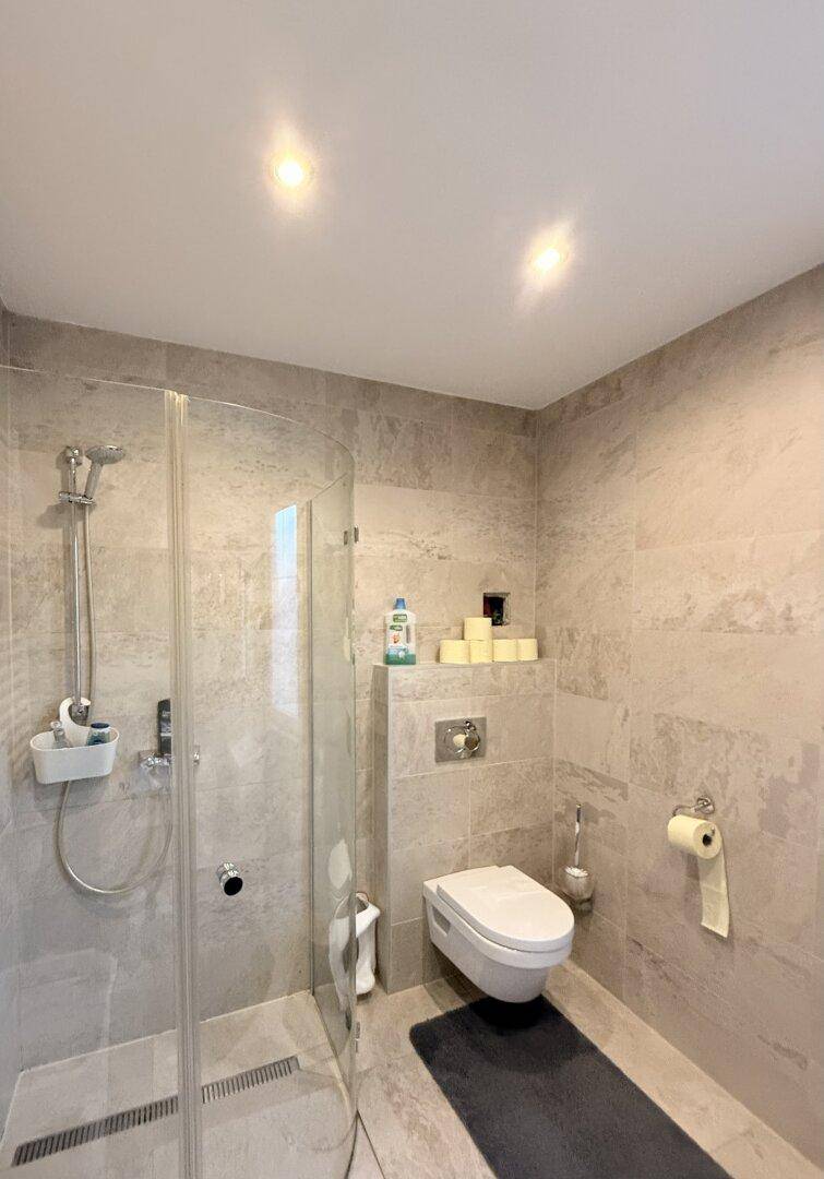 Modernes Badezimmer mit begehbarer Dusche und wandhängendem WC, in hellen Grautönen gefliest.