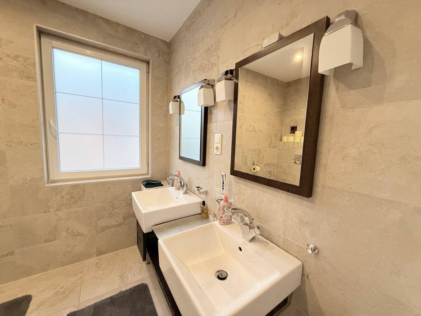 Modernes Badezimmer mit Doppelwaschbecken und großem Spiegel, in hellen Fliesen gehalten.