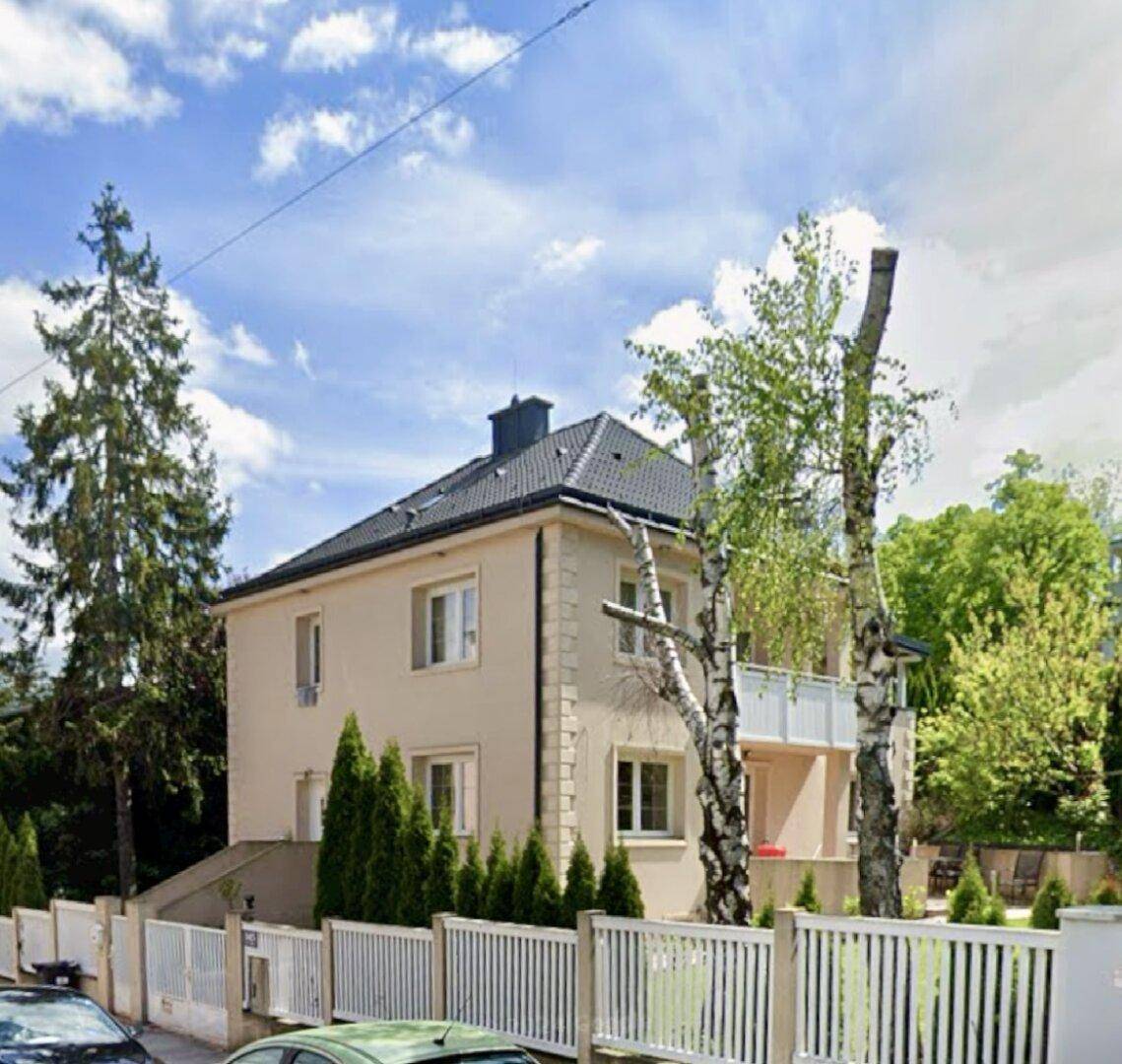 Einfamilienhaus mit Balkon und gepflegtem Vorgarten, umgeben von hohen Bäumen.