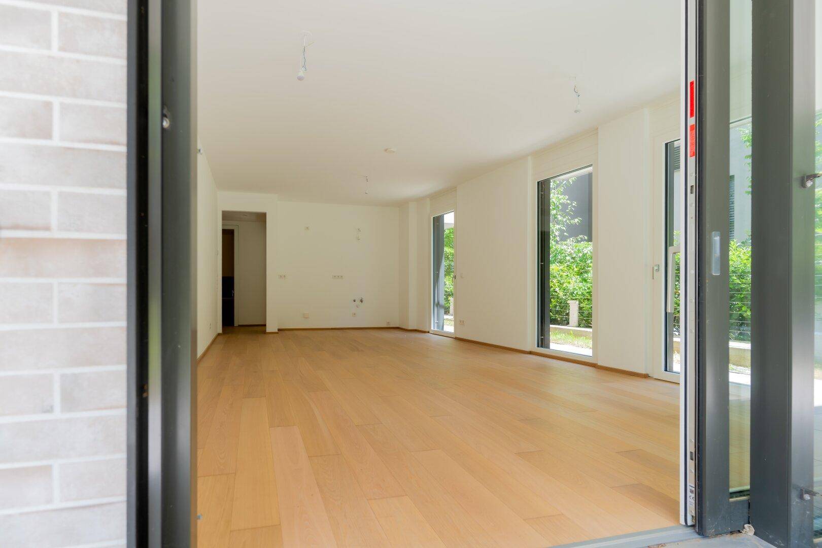 Geräumiger, heller Innenraum mit Holzboden und mehreren Fenstern zum grünen Außenbereich.