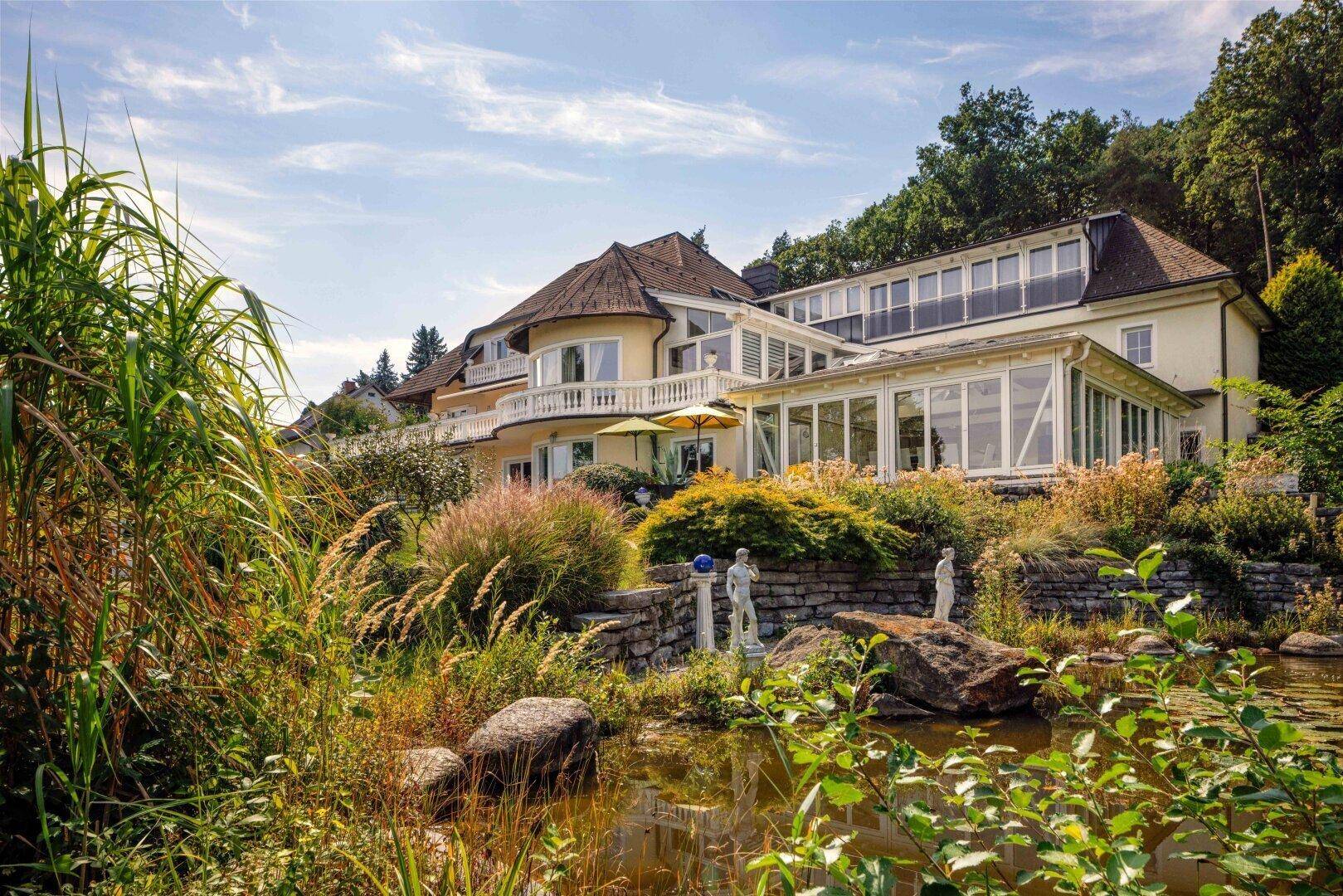 Elegante Villa mit Wintergarten und Balkonen, umgeben von einem naturnahen Teich und üppiger Vegetation.