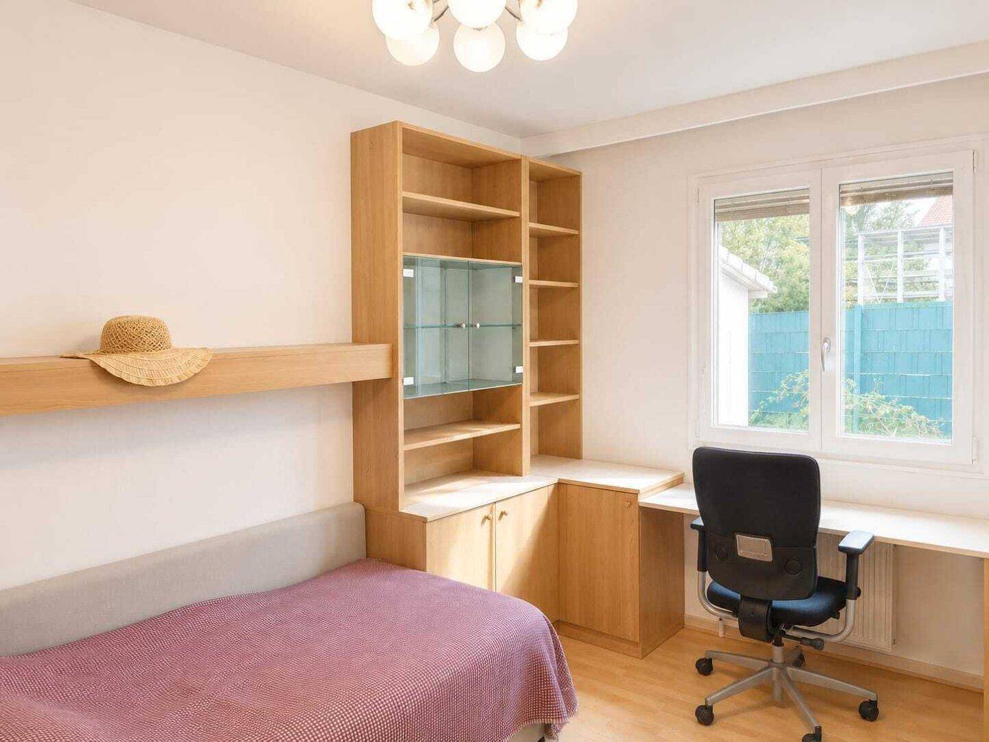 Helles Schlafzimmer mit Holzboden, Schreibtisch und Regalwand neben dem Fenster.