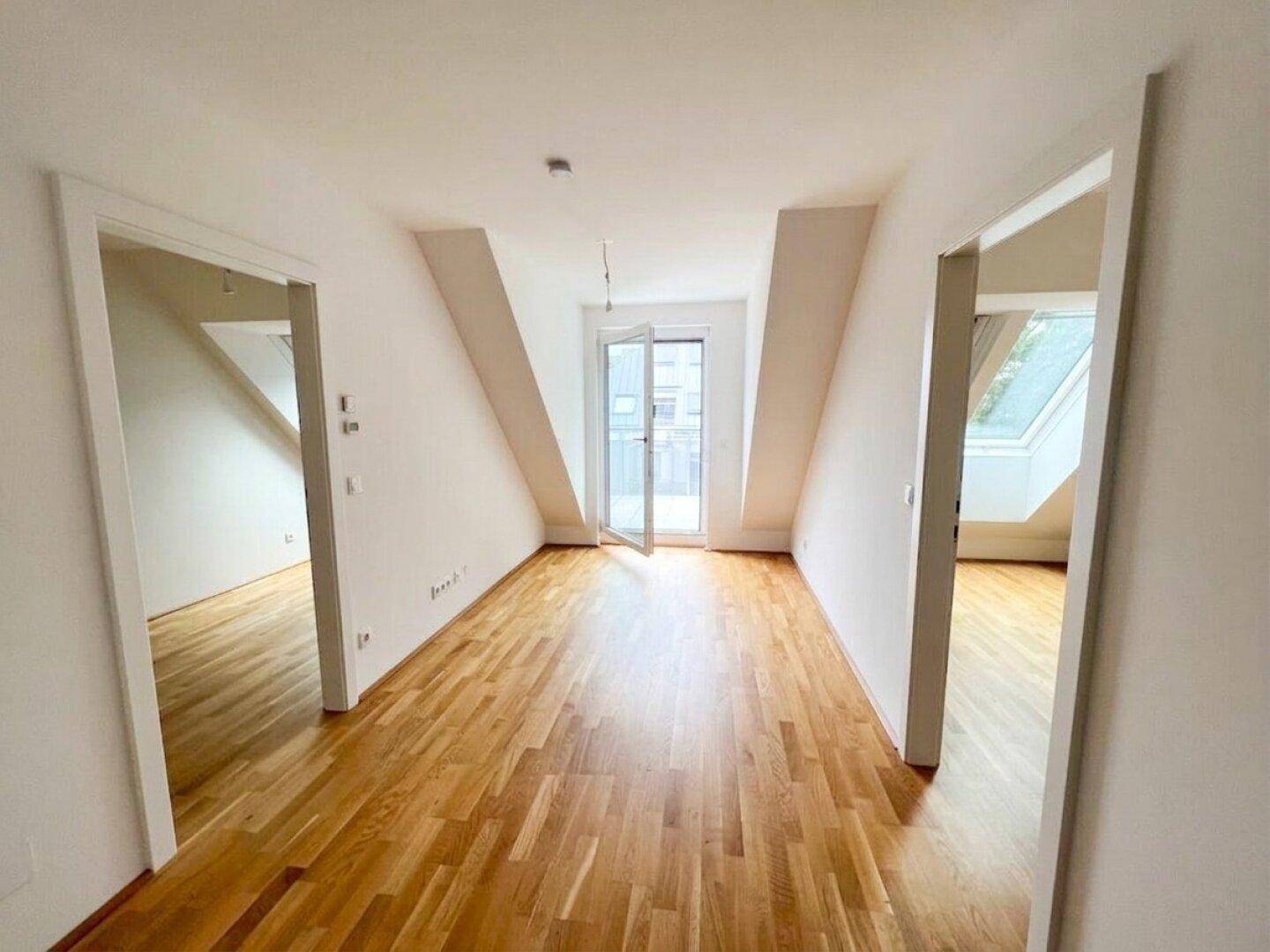 Geräumiger Wohnbereich mit Holzboden, Dachschrägen und Zugang zu einem kleinen Balkon.