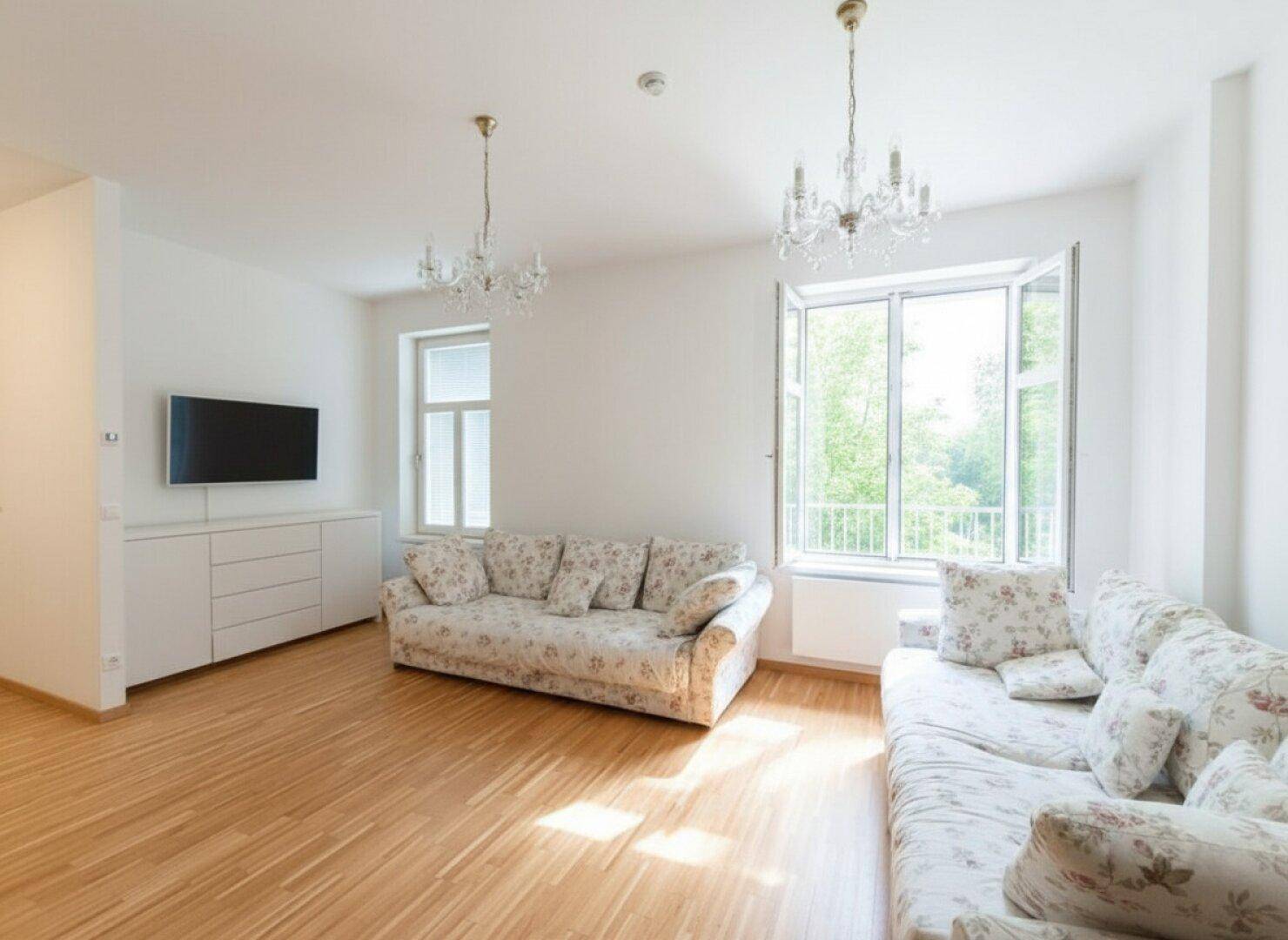 Geräumiges Wohnzimmer mit hellen Wänden, Holzboden und großen Fenstern, die viel Tageslicht hereinlassen.