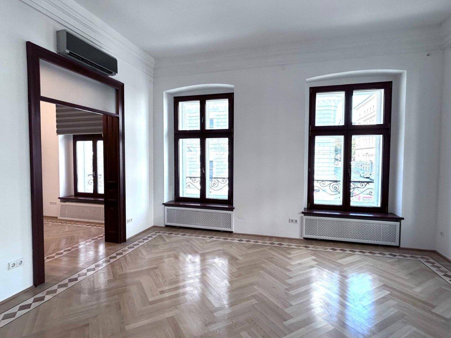 Geräumiges Wohnzimmer mit Parkettboden, hohen Fenstern und einer Klimaanlage.