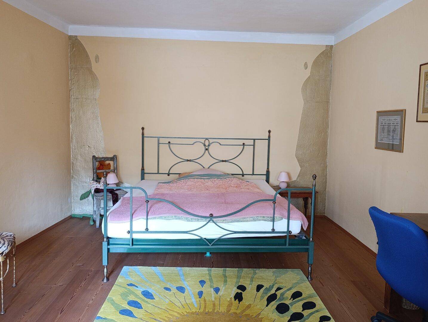 Helles Schlafzimmer mit Holzboden, Metallbett und einem auffälligen, bunten Teppich.