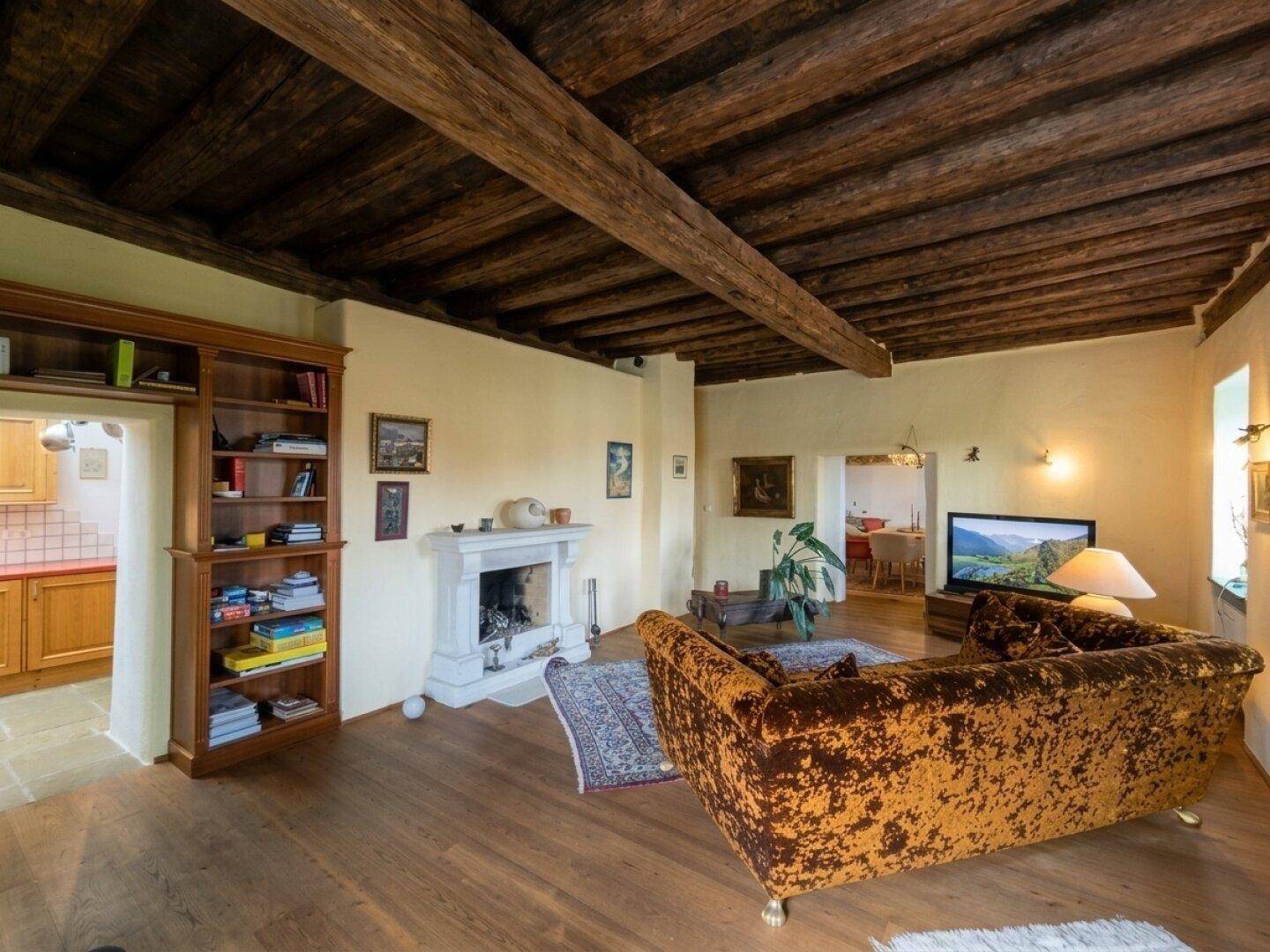 Gemütliches Wohnzimmer mit freiliegenden Holzbalken, Kamin und Blick in die Küche.