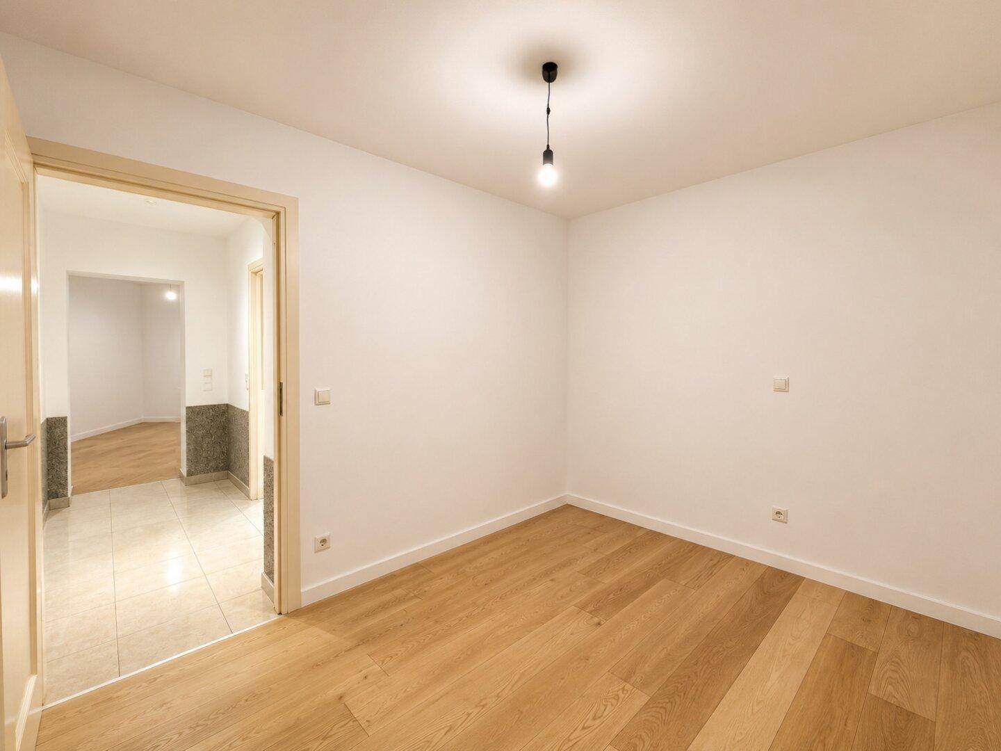 Leeres Zimmer mit Holzboden und Zugang zu einem gefliesten Flur, bereit zur Gestaltung.