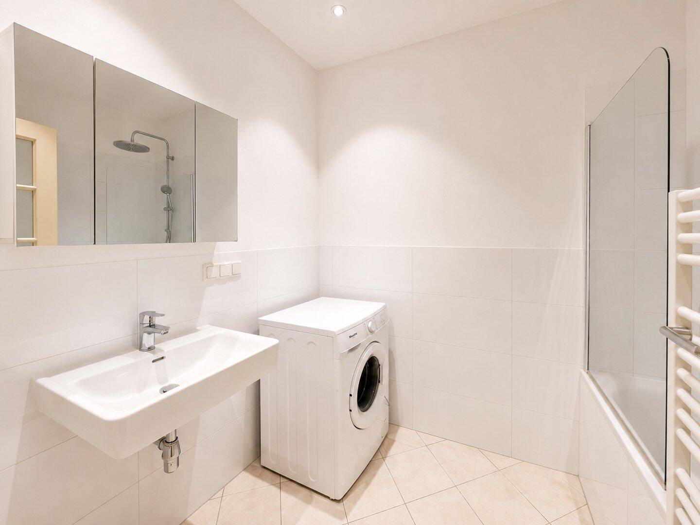 Helles Badezimmer mit Waschbecken, Spiegelschrank, Waschmaschine und Badewanne mit Duschwand.