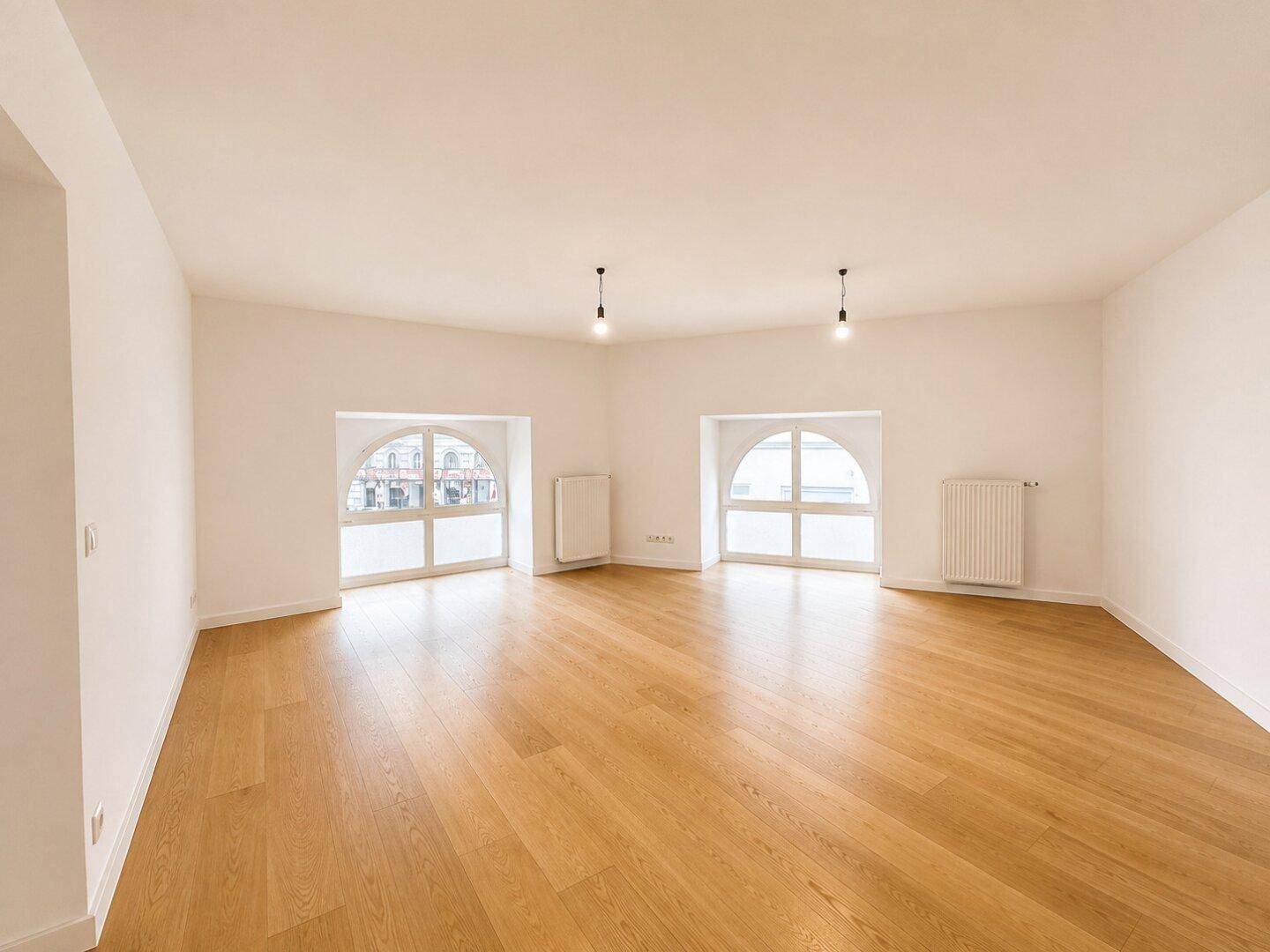 Geräumiger, unmöblierter Raum mit Holzboden und zwei Rundbogenfenstern, ideal für Wohnbereich.