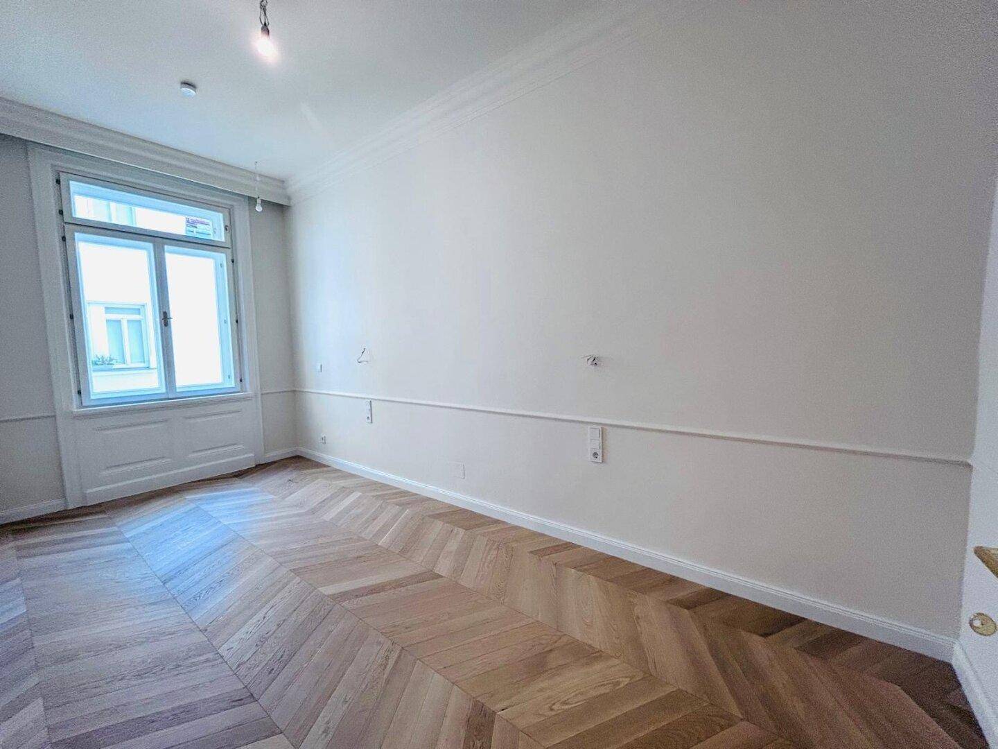 Geräumiger Raum mit Fischgrätparkett, hohen Decken und einem großen Fenster für natürliches Licht.