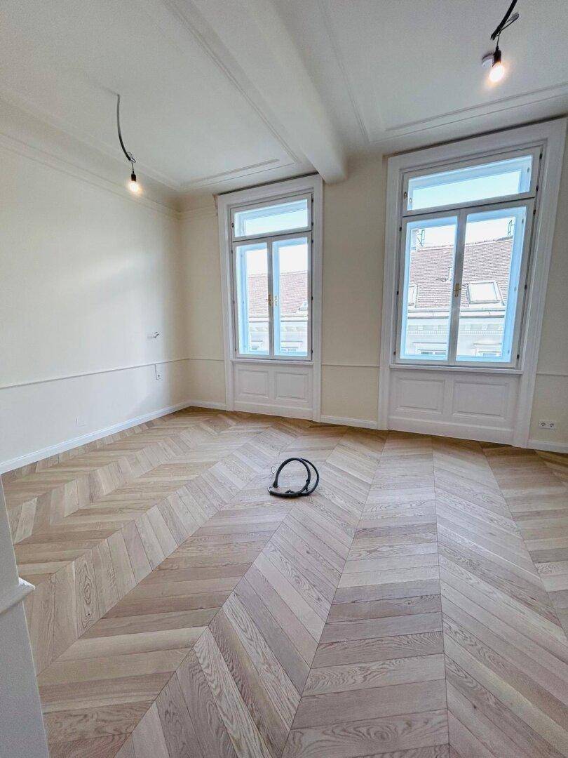 Geräumiger Raum mit Fischgrätparkett, hohen Decken und zwei großen Fenstern für viel Licht.