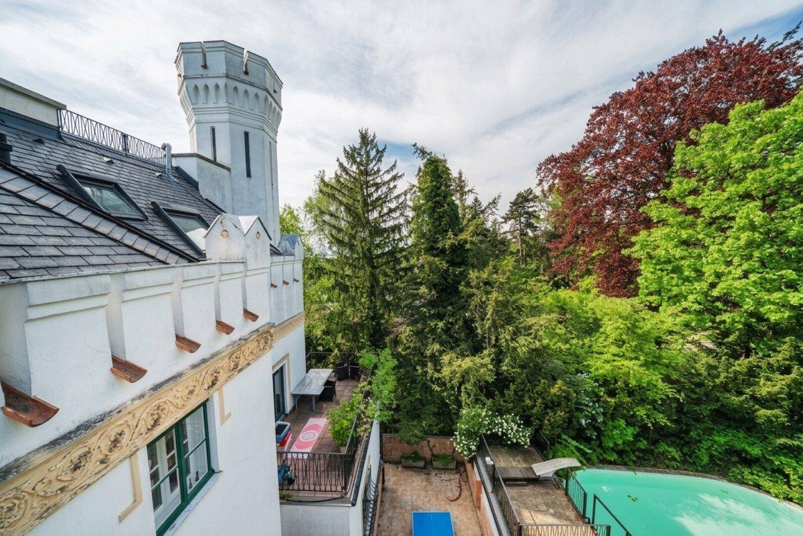Blick von oben auf das Schlossgebäude mit Turm, umgeben von dichtem Baumbestand und einem Poolbereich.