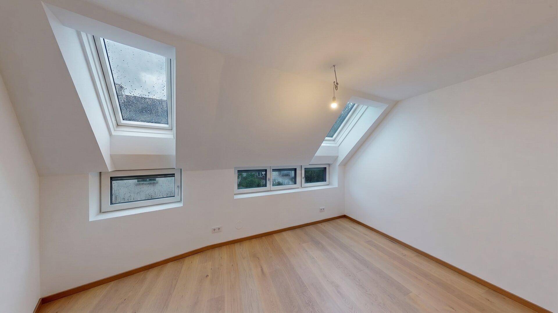 Leeres Zimmer im Dachgeschoss mit schrägen Wänden, Dachfenstern und hellem Holzfußboden.