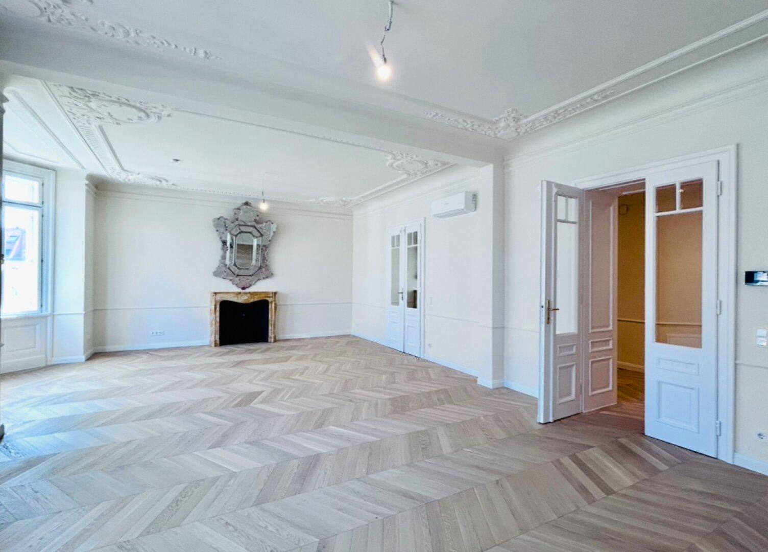 Großzügiges Wohnzimmer mit Fischgrätparkett, Stuckdecken, Kamin und Zugang zu weiteren Räumen.
