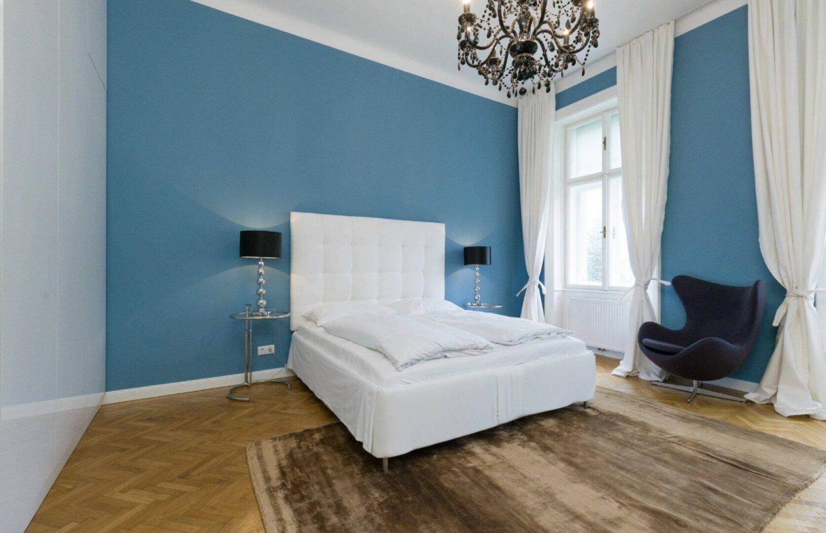Geräumiges Schlafzimmer mit blauer Akzentwand, Fischgrätparkett und einem eleganten Kronleuchter.
