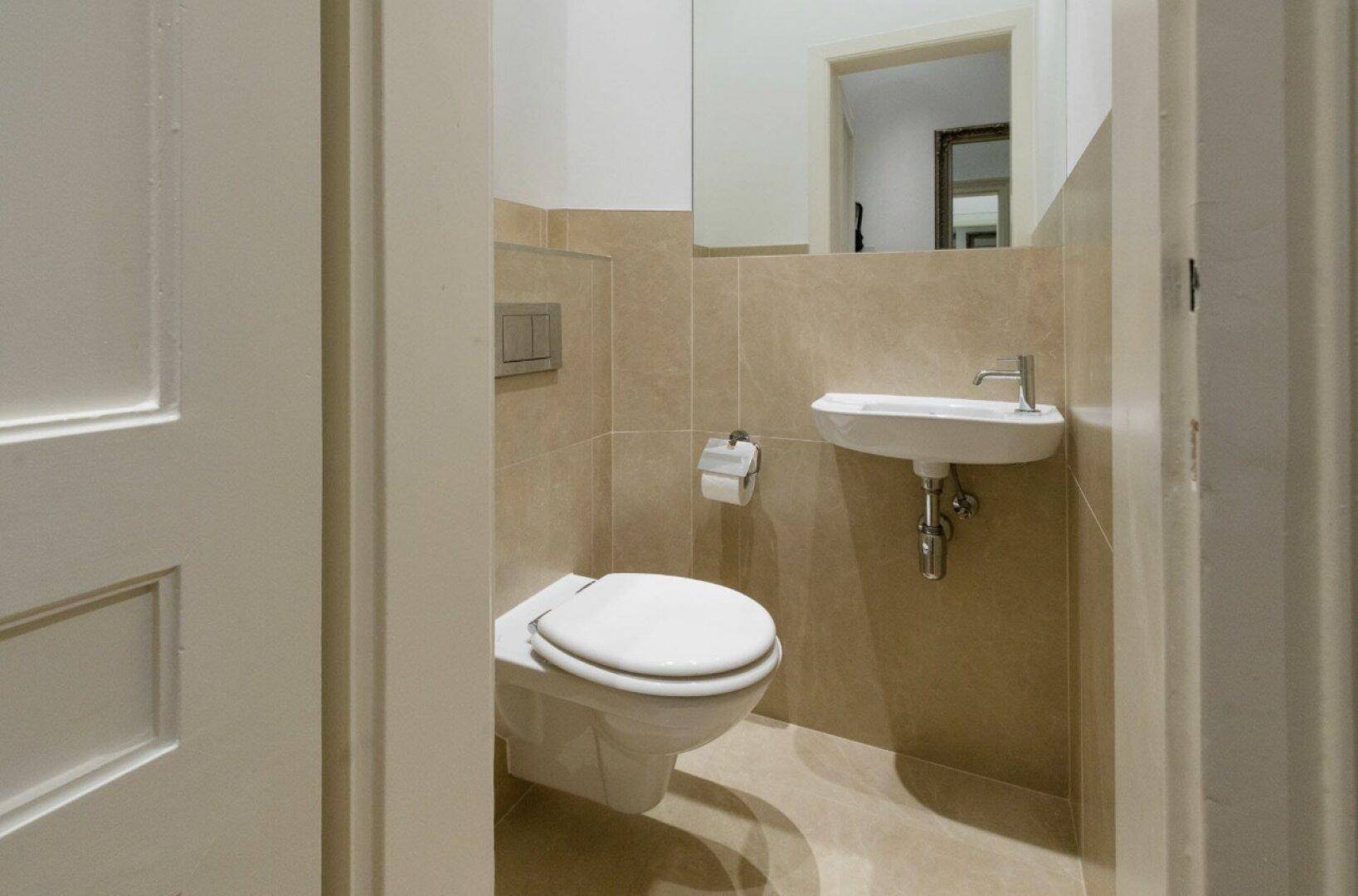 Kleines Gäste-WC mit beigen Fliesen, wandhängendem WC und einem Waschbecken.