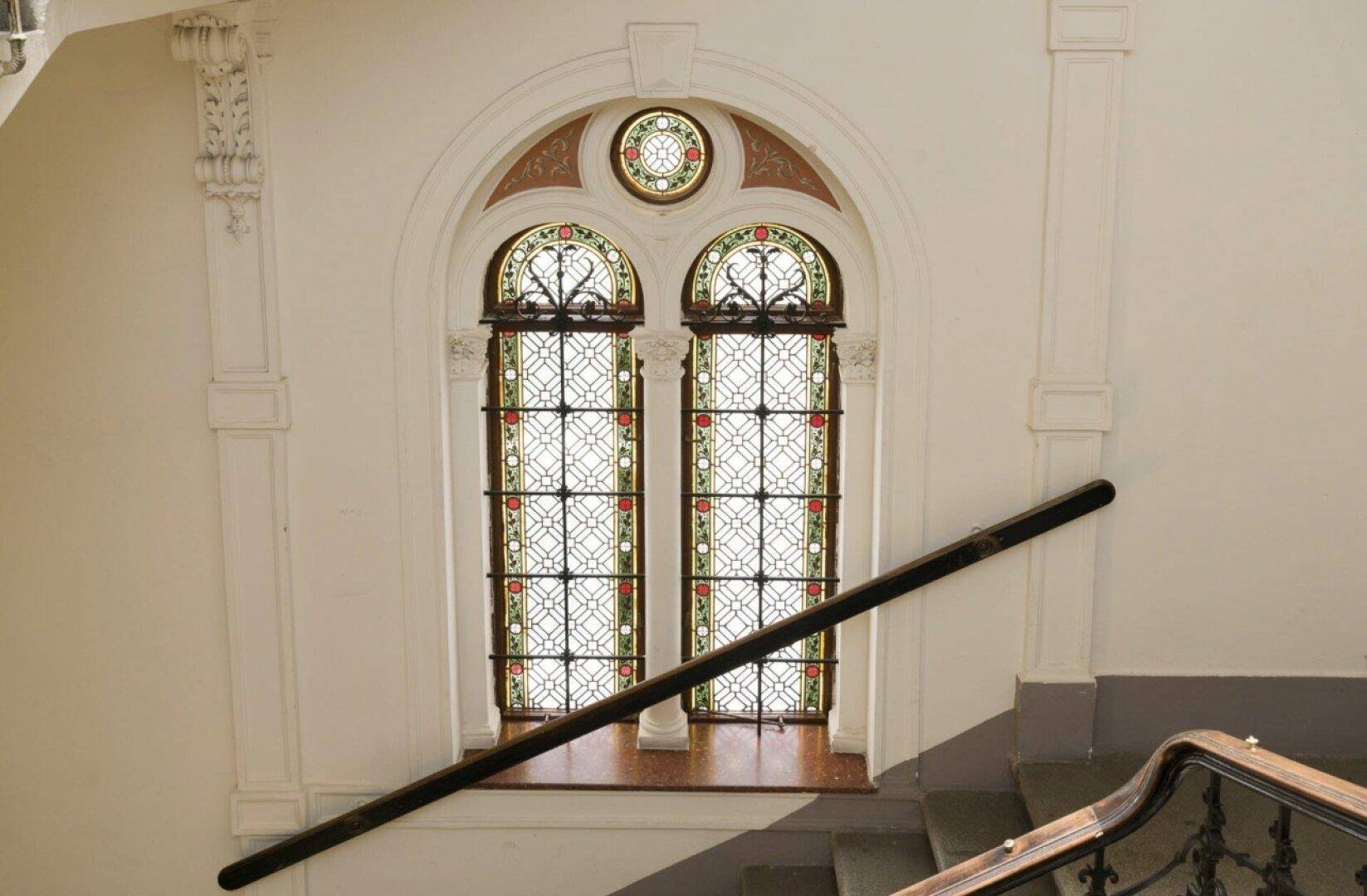 Historisches Treppenhaus mit Buntglasfenstern, kunstvollen Verzierungen und einem eleganten Handlauf.
