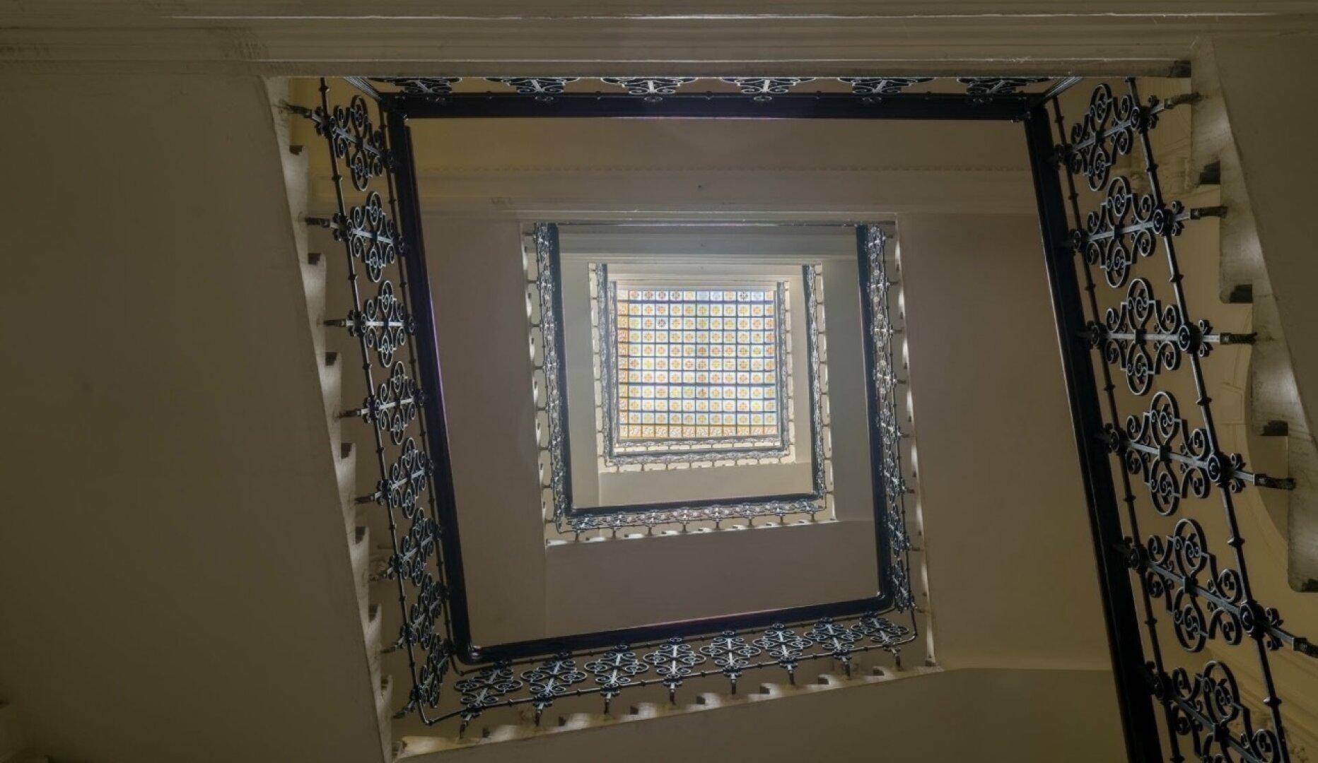 Blick nach oben in ein historisches Treppenhaus mit kunstvollem Geländer und Oberlicht.