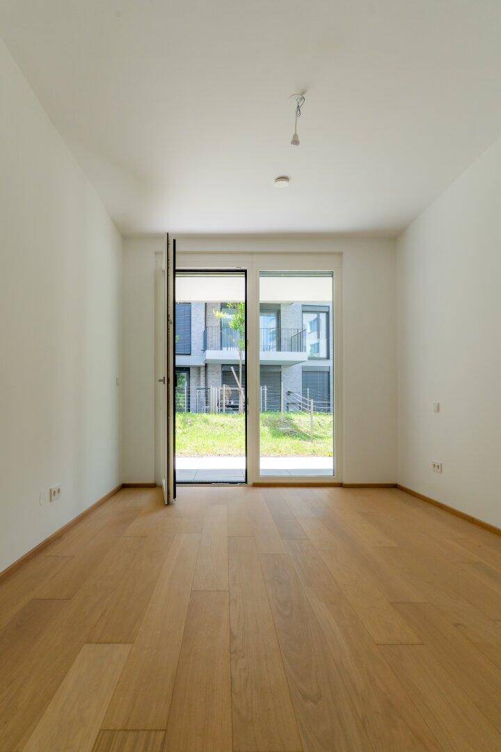 Heller, leerer Raum mit Holzdielenboden und großen Fenstern zum Außenbereich.