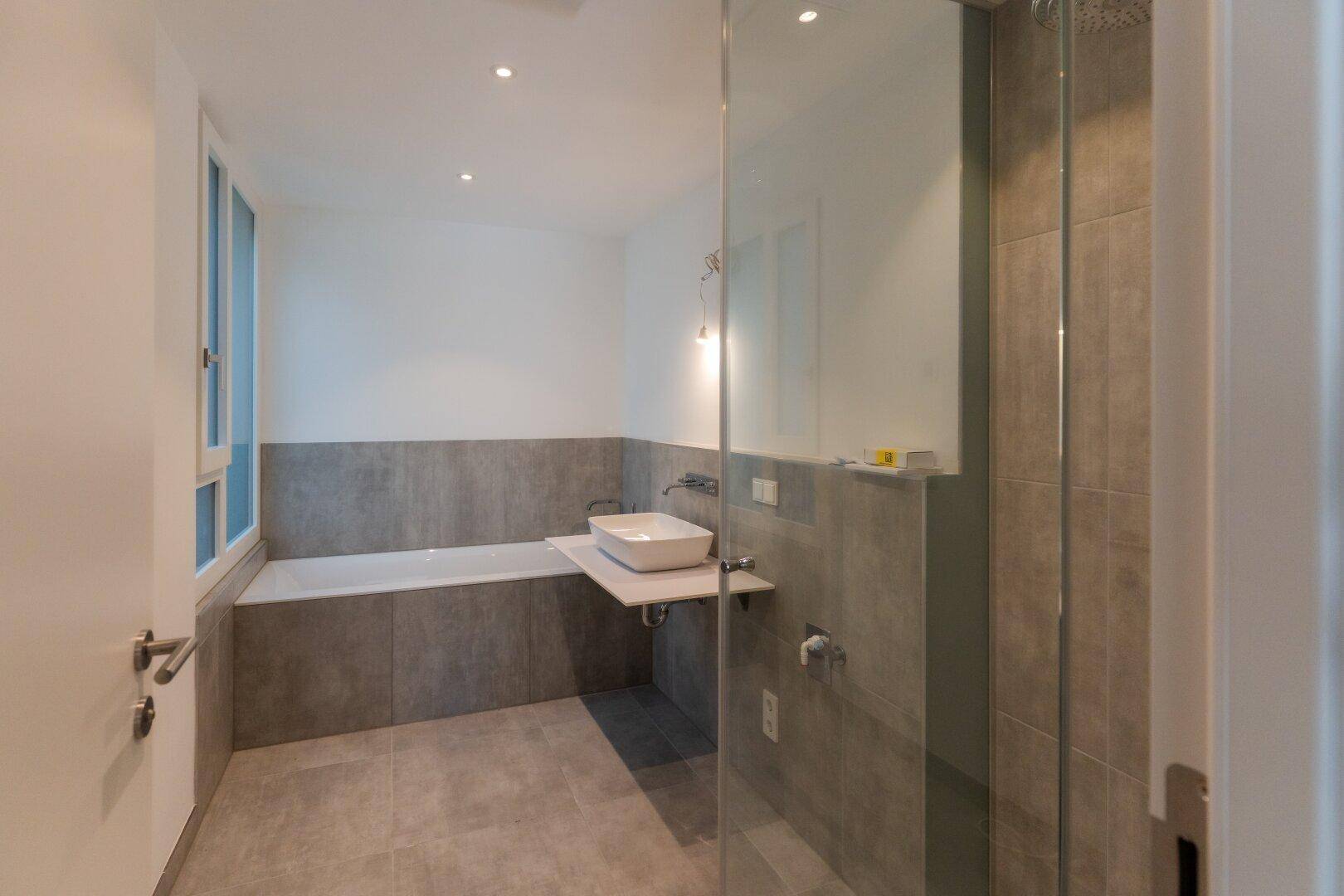 Modernes Badezimmer mit grauen Fliesen, Badewanne, begehbarer Dusche und Fenster.