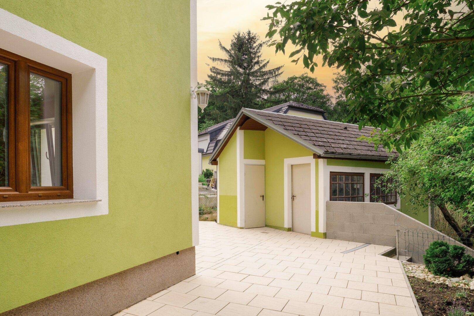 Grünes Wohnhaus mit Nebengebäude und gepflastertem Vorplatz, umgeben von Bäumen.