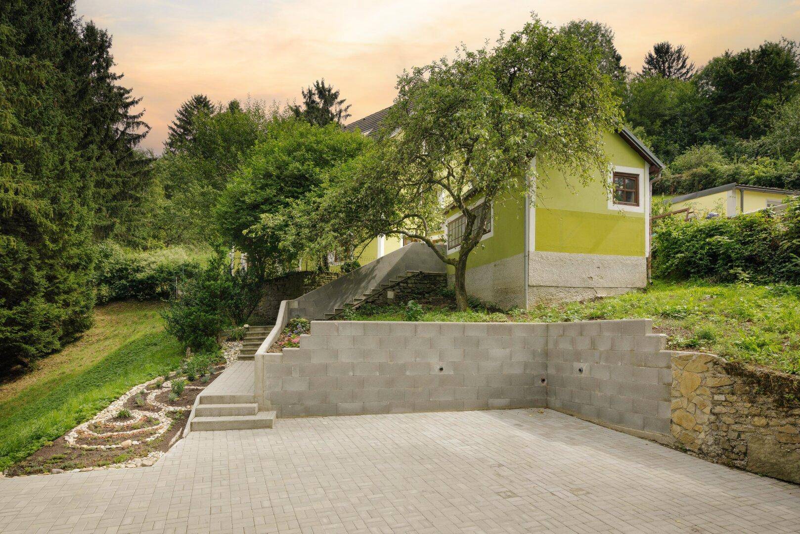 Grünes Haus auf einem Hügel mit gepflastertem Bereich und Stützmauer, umgeben von Bäumen.