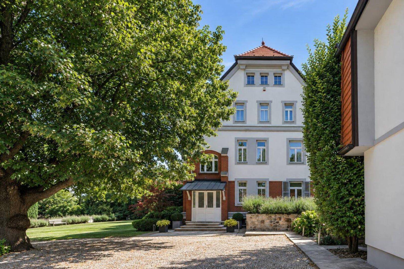 Elegante Villa mit gepflegtem Vorgarten, Kiesauffahrt und altem Baumbestand unter blauem Himmel.